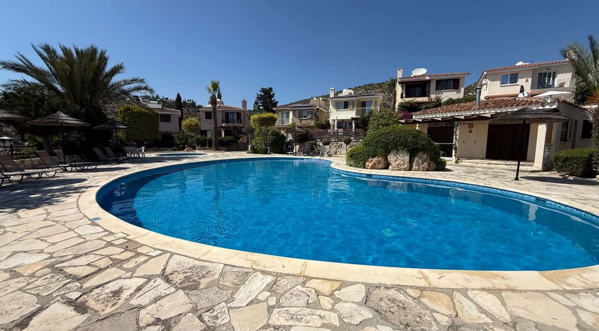 A clear blue swimming pool surrounded by stone paving, with sun loungers and palm trees nearby. Several villas with balconies and greenery are in the background under a bright blue sky.