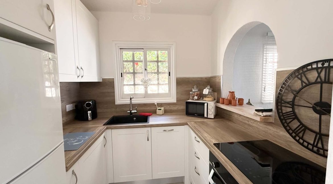 A bright, modern kitchen with white cupboards, wooden worktops, a black sink, electric cooker, microwave, jars, and a large decorative wall clock. Sunlight streams through a window above the sink.