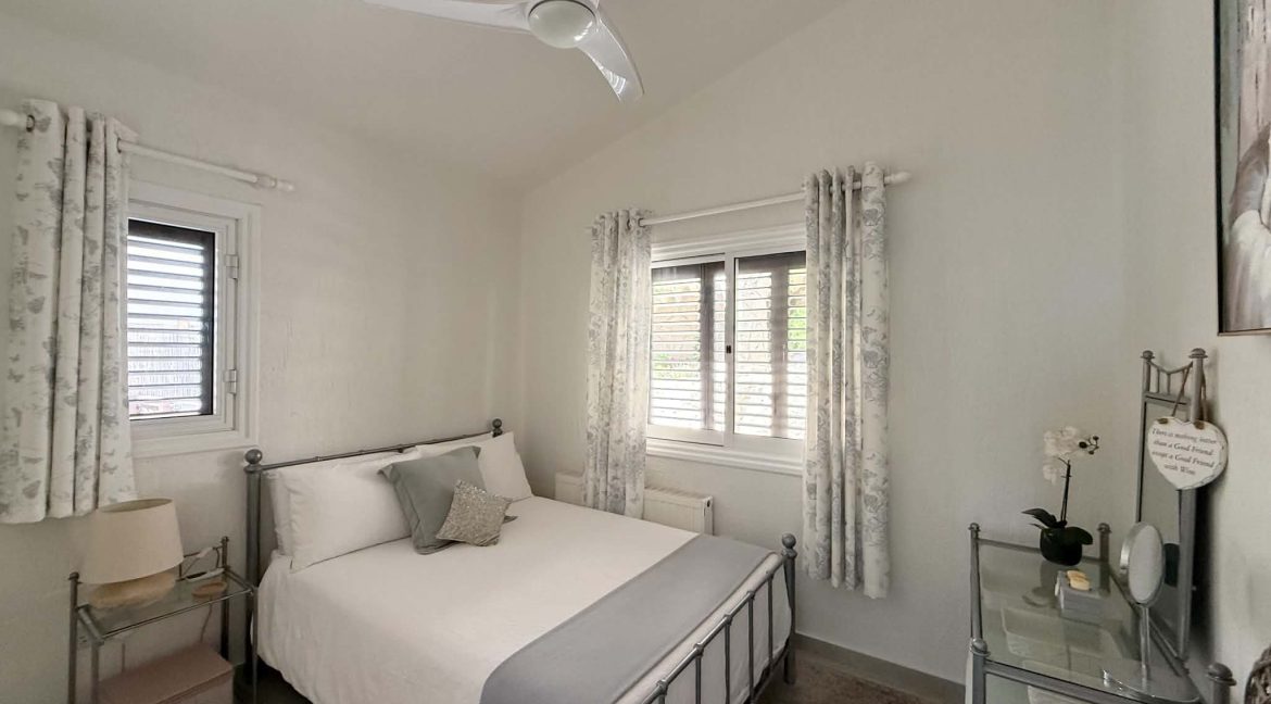 A small, bright bedroom with a single bed, white and grey bedding, two windows with floral curtains, a ceiling fan, bedside table with a lamp, and a glass desk with a chair and a potted orchid.