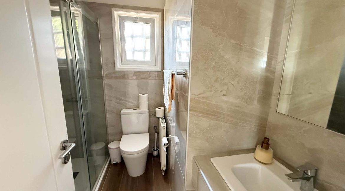 A modern, compact bathroom with beige marble walls, a glass-enclosed shower, a toilet with a toilet roll on top, a towel hanging, and a white basin with a soap dispenser next to a large mirror.