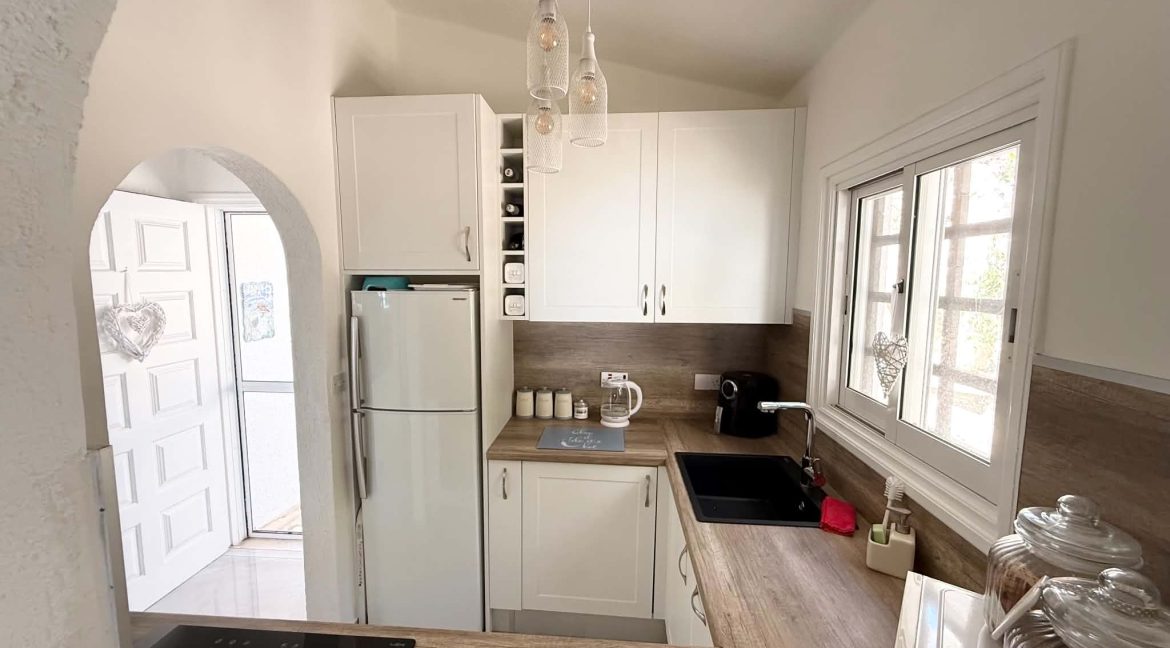 Bright, small kitchen with white cupboards, a fridge, black sink, glass jars, coffee maker, and a window letting in natural light. Three pendant lights hang from the ceiling above the wooden worktop.