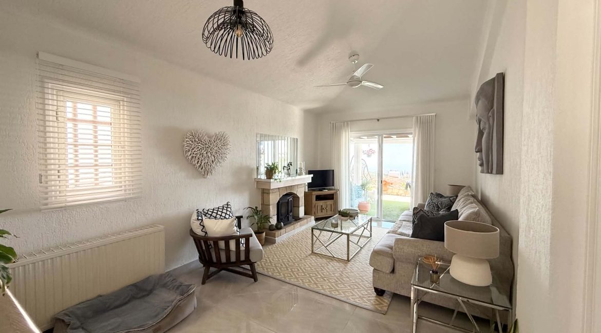 Bright living room with white walls, a fireplace, large window, and glass doors leading outside. Décor includes a grey sofa, wall art, coffee table, and wicker pet bed. Neutral tones and greenery create a cosy, airy feel.